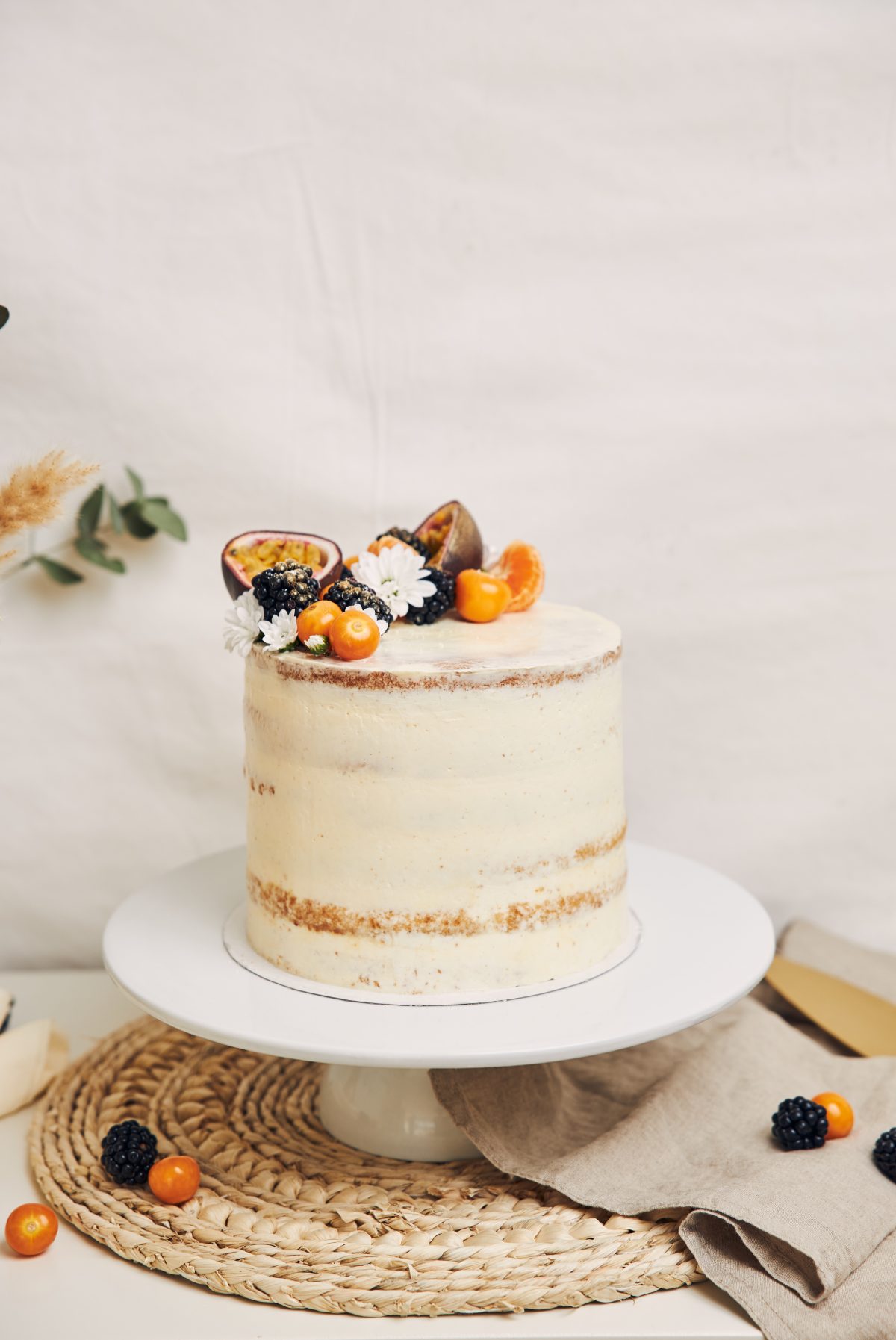 White cake with berries and passionfruits with plants behind a white background