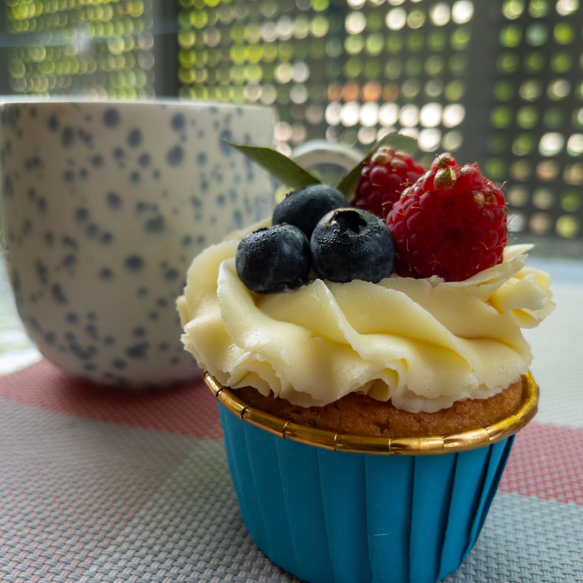 Cupcake with Berries and Coffee Mug on Outdoor Table. Delicious cupcake with cream frosting, blueberries, and raspberries