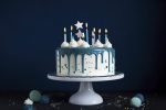White birthday drip cake with teal ganache, star toppers and fun candles over dark blue background, generate ai